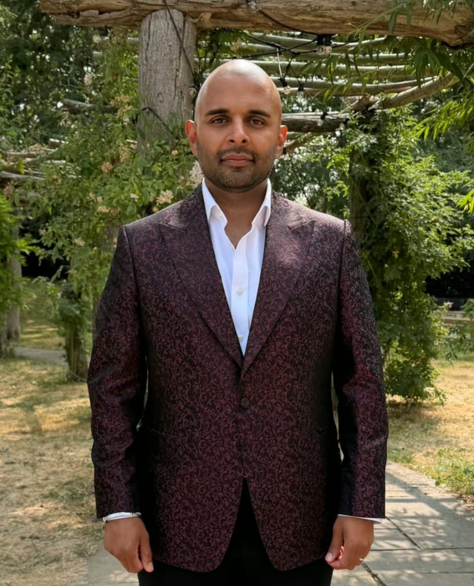 The Host Master UK in a burgundy patterned suit, photographed in a garden setting